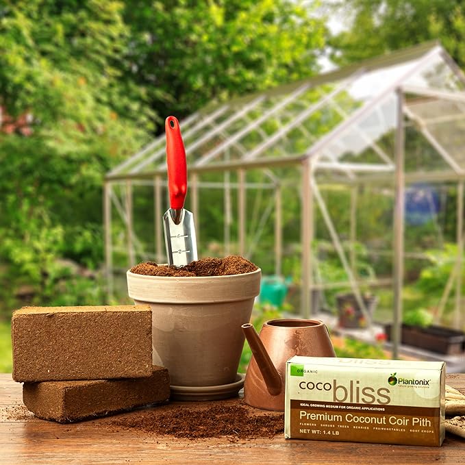 A work table featuring two open blocks of coco coir, and sealed block with the Coco bliss label, a flower pot filled with soil and a trowel, and a brass watering can. A greenhouse can be seen in the background.