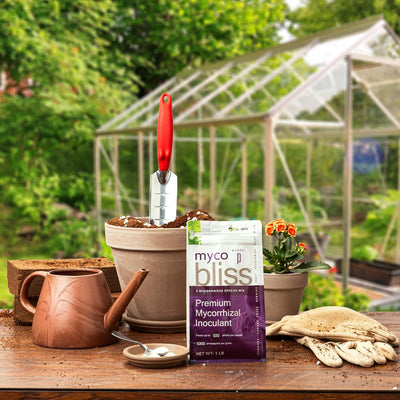 A one pound bag of premium mycorrhizal inoculant on a wooden table surrounded by various garden equipment. A green house is in the background. 